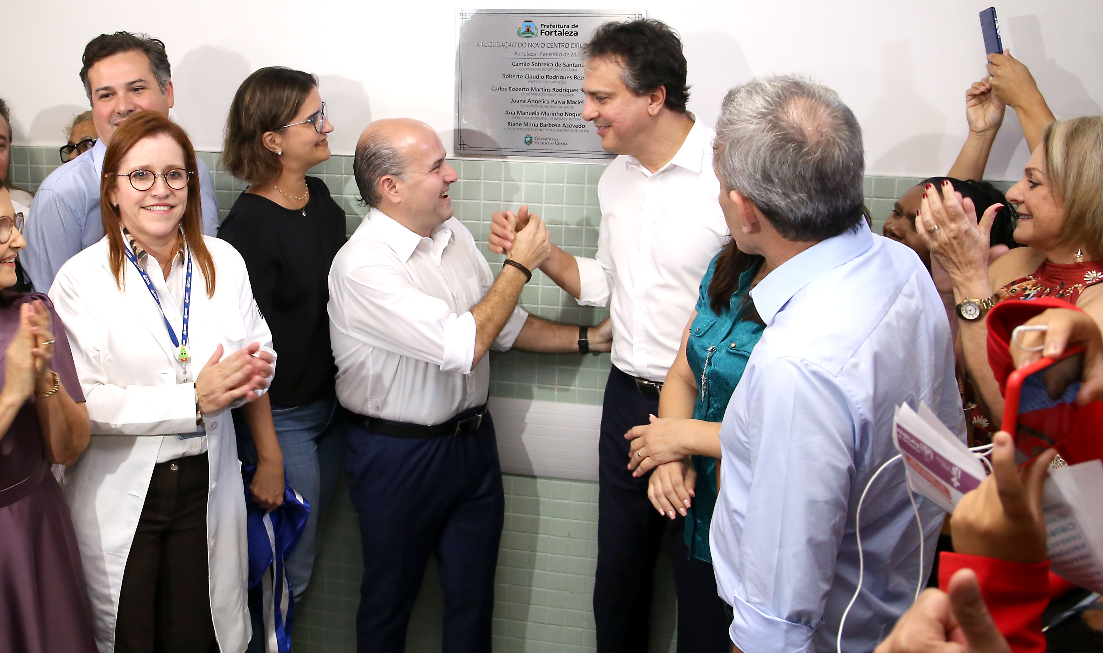 prefeito e governador sorrindo e dando as mãos em frente à placa de inauguração e pessoas aplaudindo ao redor